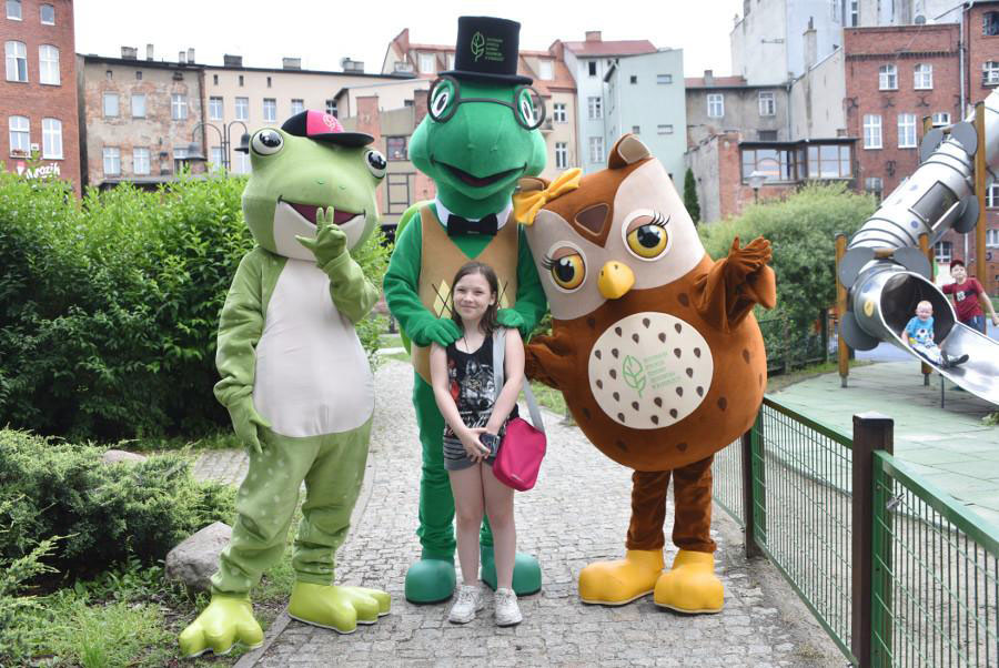 Na fotografii widnieje przebranie zielona żaba i dwie pozostałe maskotki dla RDOŚ - Żółw praz Sowa. Pozują do fotografii z dziewczynką, obok widnieje plac zabaw dla dzieci. W tle zabudowania miasta. 